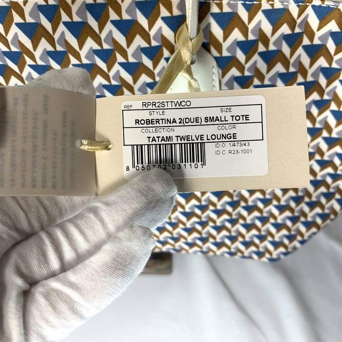 Bàn tay đeo găng tay cầm thẻ sản phẩm "ROBERTINA 2 (DUE) SMALL TOTE" bên trên lớp vải họa tiết tatami với hoa văn hình học với màu xanh lam, nâu và trắng, là phong cách đặc trưng của bộ sưu tập tote nhỏ của ROBERTA PIERI.