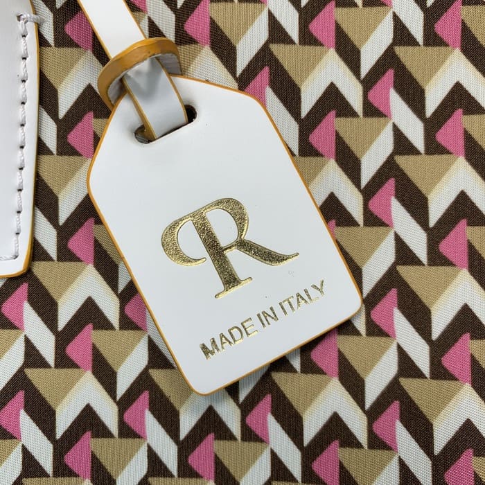 Cận cảnh thẻ hành lý Roberta Pieri màu trắng với chữ "R" bằng vàng và dòng chữ "MADE IN ITALY" được gắn vào một chiếc túi vải thô mini có hoa văn hình học màu nâu, be và hồng.