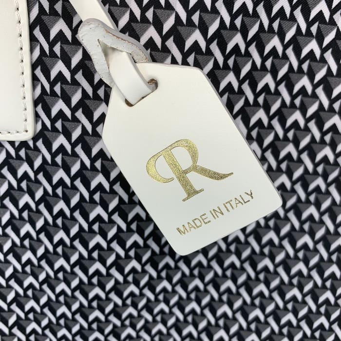 Thẻ ROBERTA PIERI bằng da trắng với các chữ cái "R" bằng vàng và dòng chữ "MADE IN ITALY" treo trên nền của một loại vải hình học đen trắng.