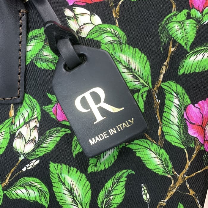 Thẻ hành lý ROBERTA PIERI màu đen với chữ "R" màu vàng và chữ Ý được treo trên vải hoa với lá xanh và hoa hồng, bổ sung cho chiếc vải thô mini với sự sang trọng vượt thời gian của Ý.